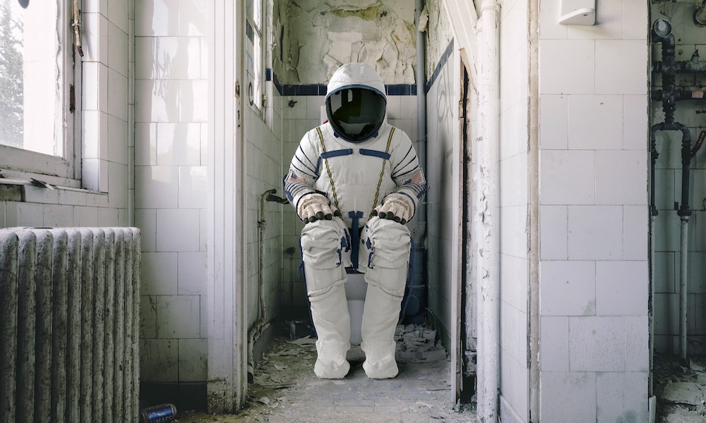 An astronaut sits on a toilet in a run down white tiled bathroom