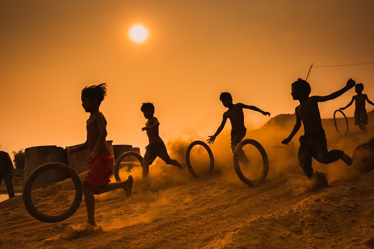 A group of children with bike tires running down a sandy hill keeping the tires rolling.