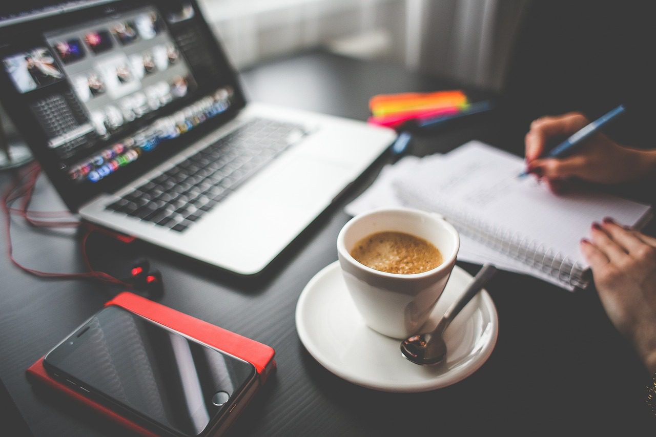 A table with a laptop, cup of coffee, a phone, and a person writing in a notebook