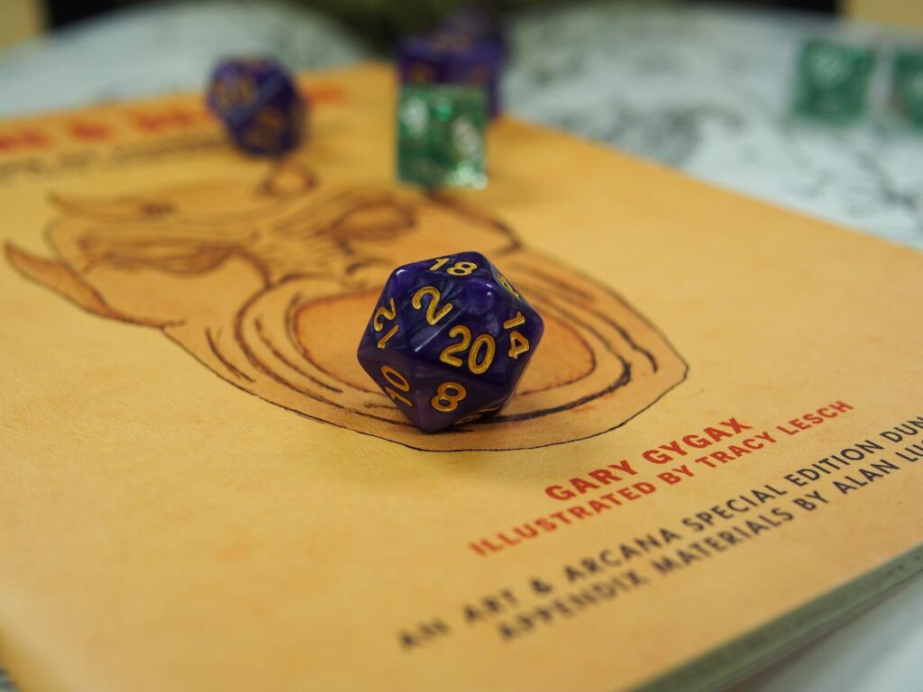 Close-up of a Dungeons & Dragons book featuring artwork and the name Gary Gygax. A purple twenty-sided die (d20) with gold numbers, showing a 20, rests on top of the book.