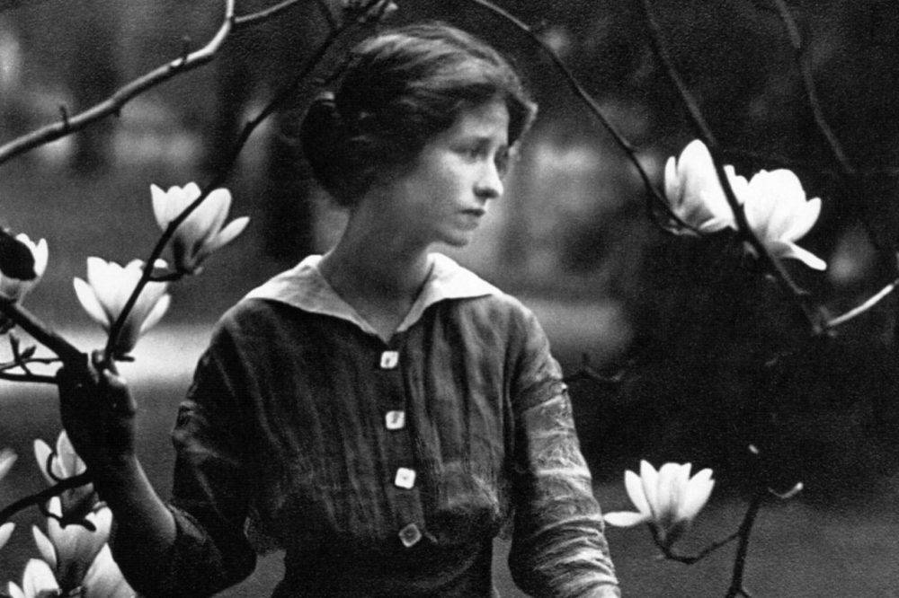 image of poet Edna St. Vincent Millay. black and white image of a woman standing and looking to the right with branches of a flowering tree in the foreground