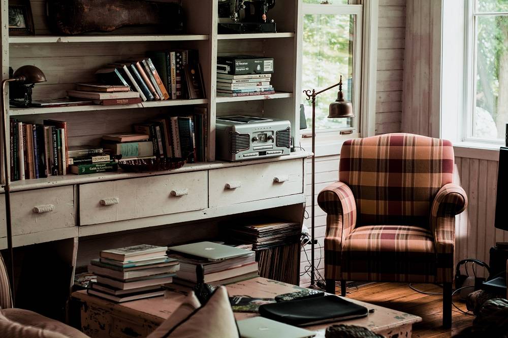 a cozy reading room with natural lighting, a plaid armchair, and stacks of books