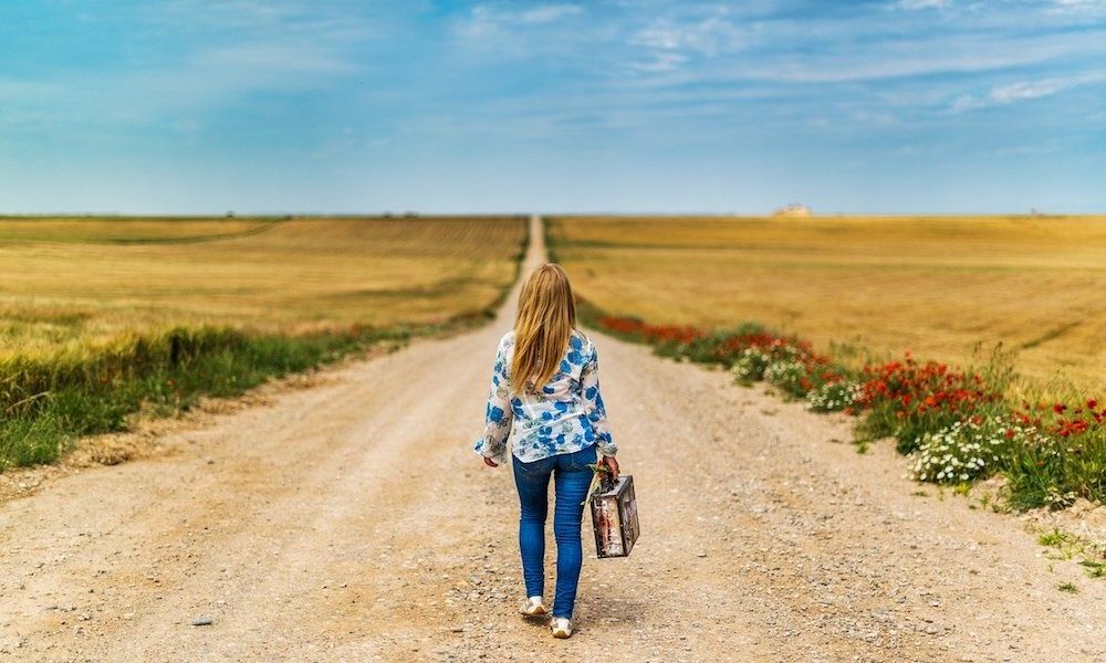 a girl walks down a dirt road with a suitcase with her back to the camera